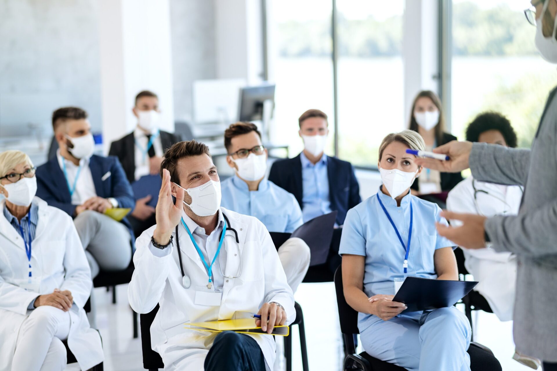 Happy doctor with protective face mask raising hand to ask a question while attending educational event.